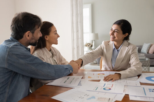 Smiling professional Indian woman consultant in formal attire shakes hands with couple across desk filled with financial charts, graphs, and reports. Successful conclusion of consultation or agreement