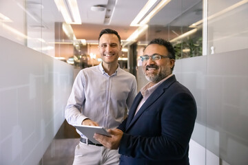 Two happy diverse young and senior coworkers posing for professional portrait in office corridor, using modern technology on digital tablet, standing together, looking at camera, smiling
