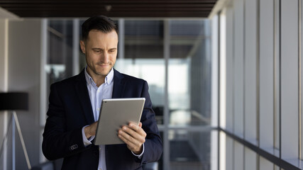 Fototapeta premium Positive successful Latin business professional man typing on digital tablet, using financial service on gadget for investment management, browsing social media. Banner shot with copy space