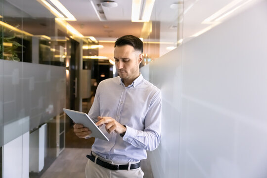 Serious successful Hispanic businessman using Internet technology on tablet for work online communication, standing in office corridor, leaning on glass wall, typing on digital gadget. - Powered by Adobe