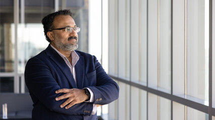 Positive pensive senior Indian company executive man looking at large office window, standing in...