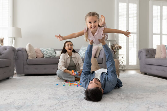 Happy father lying on rug, holding his little 3s cute daughter up in air with hands stretched during family pastime in modern living room. Playtime activity, playful interaction between parent and kid