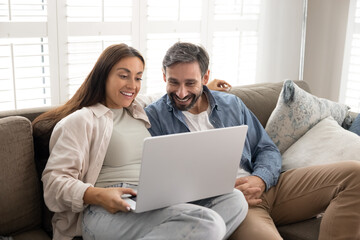 Happy young couple sitting on comfortable couch in living room, browsing internet, buying goods and electronic services together, watching movie or TV show using modern laptop. Marketplace, e-commerce
