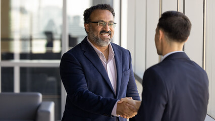 Cheerful older Indian business owner man shaking hands with younger Caucasian consultant, job candidate, smiling, laughing, giving greeting handshake to business partner. Banner shot © fizkes