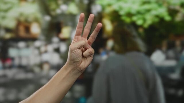 Man holding three fingers on a sunny city street with blurred backgrounds showing trees and people.
