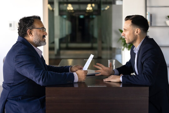 Fototapeta Two diverse male younger and older business coworkers arguing at work table, sitting opposite, talking, having conflict of generations. Senior boss dissatisfied with young professional paper report