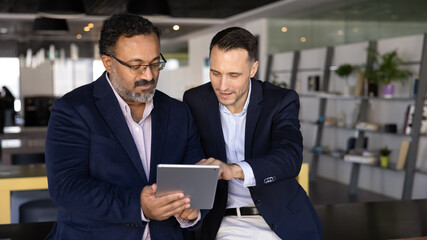 Two focused diverse young and mature businessmen browsing professional social media on tablet. Younger employee giving help to elder colleague with online application work. Banner shot