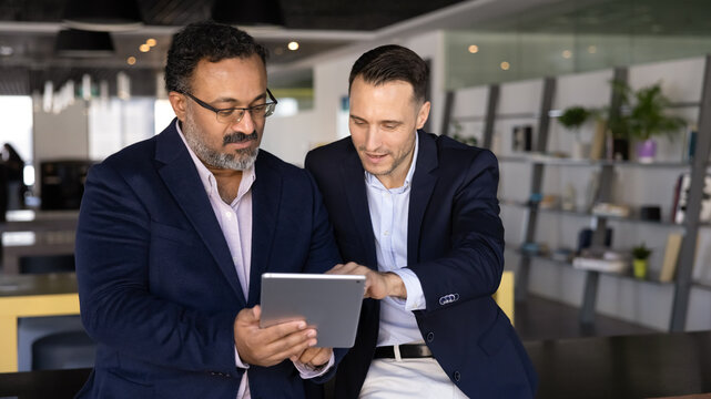Two focused diverse young and mature businessmen browsing professional social media on tablet. Younger employee giving help to elder colleague with online application work. Banner shot
