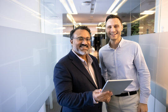 Happy diverse elder and younger male business partners standing together in office corridor, posing for professional teamwork portrait, holding digital tablet, looking at camera, smiling - Powered by Adobe