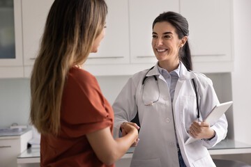 Cheerful young Latin practitioner woman shaking hands with female patient, congratulating on...