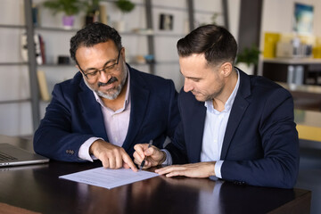 Senior Indian businessman pointing finger at place for signature in document for younger business partner signing. Real estate, insurance agent, notary meeting with client