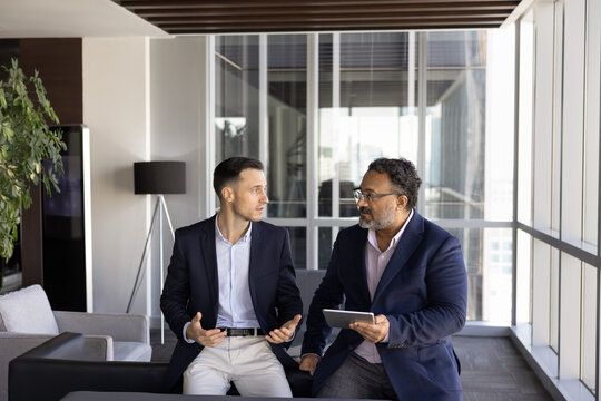 Two younger and elder diverse male business colleagues talking in office lobby, discussing work project, teamwork, collaboration plan, speaking, holding tablet. Businessman consulting coworker