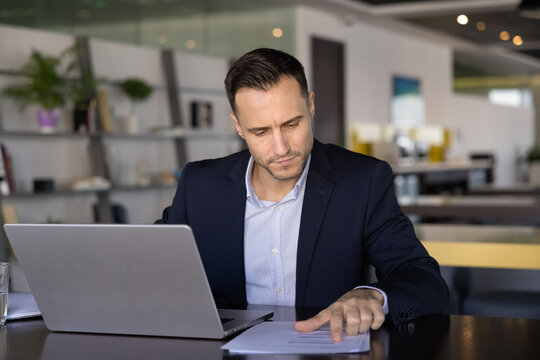 Focused Latin business product owner man analyzing paper sales marketing report at office workplace, sitting at laptop, pointing at document, working on project strategy
