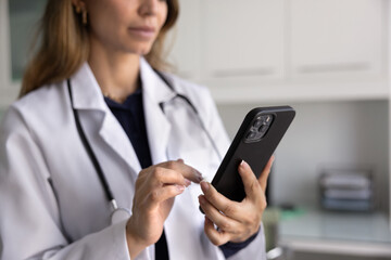 Close up of smartphone in hands of young female doctor typing message to patient, using digital gadget and web service for telemedicine, online consultation. Cropped shot
