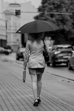 Fototapeta Black and white photo of businesswoman walking with umbrella, professional woman in skirt and blazer holding bag, rainy city commute, urban lifestyle, stylish pedestrian in monochrome street scene
