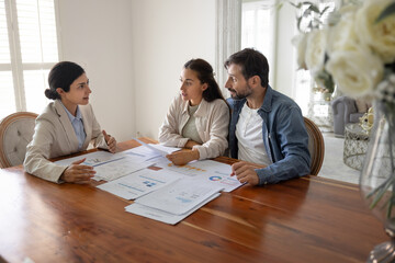 Professional consultant meets with couple at table covered with documents, graphs, and data charts, explains points, specific sections, review detailed plans or reports. Budgeting, financial planning