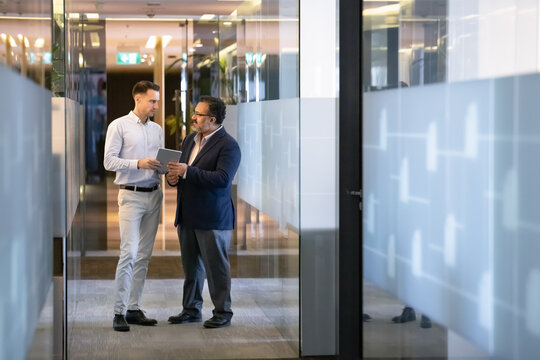 Elder Indian boss and younger manager discussing work on online project, using digital tablet together, standing in office corridor with glass walls, talking. Vertical full length shot