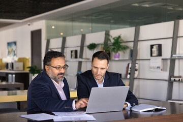 Two busy young and elder male business colleagues sharing laptop at workplace, using computer for...