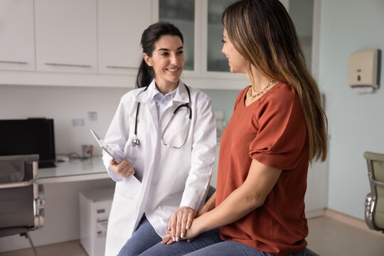 Happy optimistic young Latin physician meeting with patient after examination, touching hands of woman in supportive gesture, telling good news from medical report, smiling