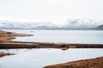 A distant car drives across a lagoon in Snæfellsnes, Iceland, with the sea and snowy fjord in the...