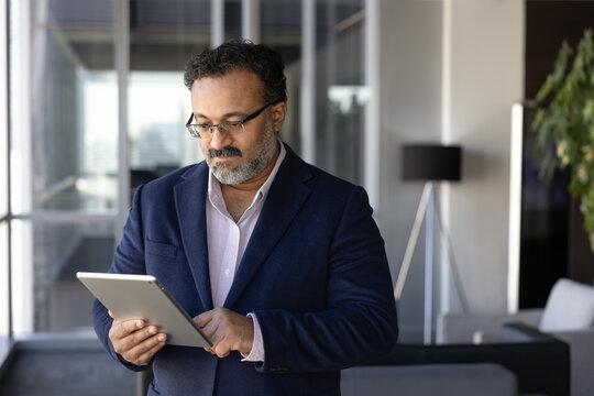 Fototapeta Serious Indian company director man using management application, online trading service, financial app on digital tablet for business communication, standing in office or hotel lobby