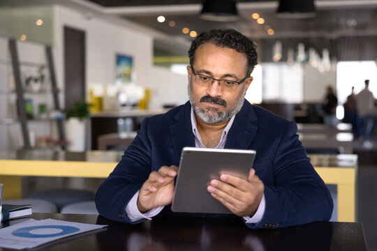 Serious elder Indian executive man typing on tablet at table in open office co-working space, using online communication technology for marketing analysis job, reading, watching content