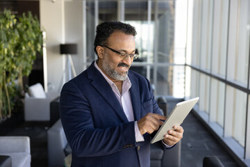 Fototapeta premium Positive older male Indian CEO using online financial web service on digital tablet in office lobby, enjoying modern Internet technology, solution for business, communication