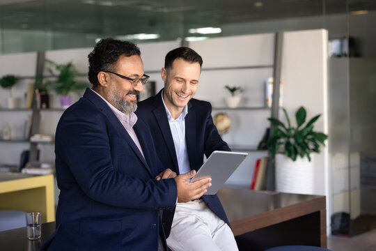 Two happy diverse male mature and older colleagues using tablet computer on work break, watching online professional content on digital gadget, laughing, enjoying Internet communication