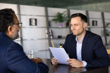Younger employer man reading resume of senior candidate with doubts on job interview. Mature Indian applicant getting rejection from human resource manager, staying unemployed