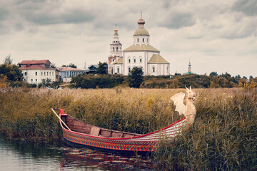 Ancient Russian church with grassy river bank and historic wooden boat on cloudy day, Suzdal, Russia