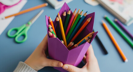 Child holding a purple paper container full of colored pencils on a blue surface with scissors