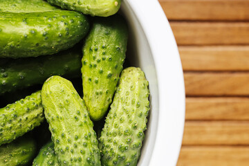 Fresh green cucumbers in a white bowl on wooden table background. Organic vegetables with textured skin ready for cooking or pickling. Healthy natural food concept with rustic kitchen style.