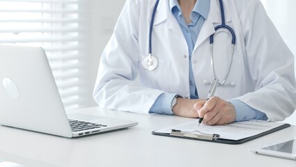 Female doctor documenting patient information while working with digital devices in a minimalist white workspace. Medicine and health care concept