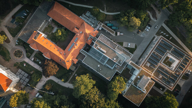 Aerial top-down view of a building complex with a red brick roof and modern structures with solar panels, surrounded by trees and green areas, captured in the evening light.