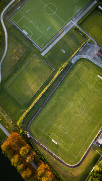 Vertical aerial top-down view of sports fields and running track surrounded by green grass and autumn-colored trees at sunrise.