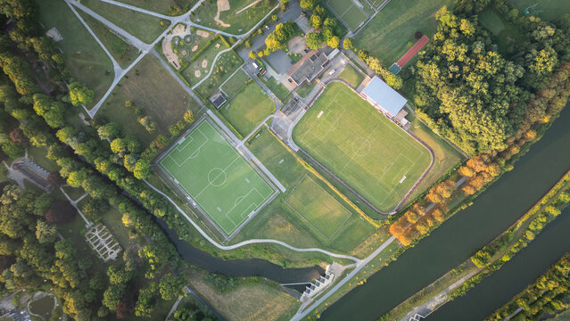 Aerial drone view of a sports complex with multiple football fields, running tracks, and green areas located along a canal.