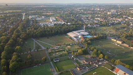 Aerial view of a suburban area with residential houses, green fields, and a canal at sunrise, captured by drone.