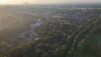 Aerial view of a city surrounded by dense green forest at sunrise, with soft morning light and mist in the distance, highlighting urban and natural landscapes.