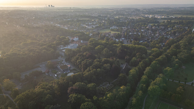 Aerial view of a city surrounded by dense green forest at sunrise, with soft morning light and mist in the distance, highlighting urban and natural landscapes.