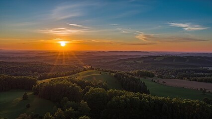 Fototapeta premium Tranquil summer evening scene from above, displaying verdant fields, scattered trees, and a bright orange sun rising over distant hills with copy space.