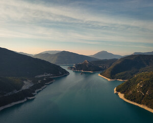 Aerial view of a serene lake winding between forested mountains under a soft pastel sky at sunset.
