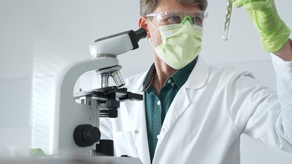 Masked senior man researcher examining green botanical samples in test tube in laboratory during...
