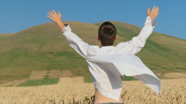 Young attractive man in unbuttoned white shirt stands in ripe wheat field facing mountains, back to camera, arms spread wide, celebrating life, freedom, success, and pure happiness.