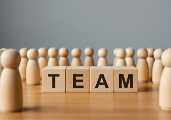 A group of wooden peg doll figures stands together around blocks spelling "TEAM," representing business teamwork, collaboration, unity, and a strong company community.