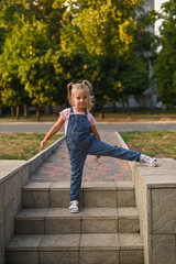 girl in denim jumpsuit is standing on steps outside with one leg stretched out to side. In background are green trees in soft evening light. girl in jumpsuit poses on stairs against backdrop of park.