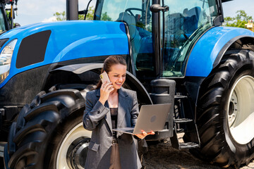 Businesswoman with laptop and mobile phone standing in front of new tractor at agricultural...