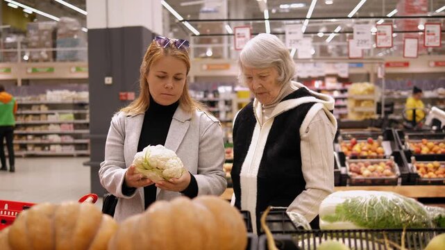 An adult daughter helps her grandmother choose fresh cauliflower in a large supermarket, they stand near a shelf with vegetables and talk. Healthy nutrition for the elderly.