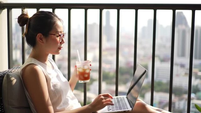 Asian woman enjoys remote work with a laptop and iced tea on a modern city apartment balcony overlooking the skyline.