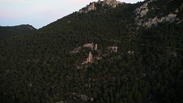 Aerial drone video of a forest-covered mountain peak in Spain at sunrise, with warm sunlight casting golden tones on rocky slopes.