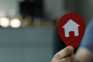 A hand holds a red location marker with a house symbol, representing finding the perfect home.